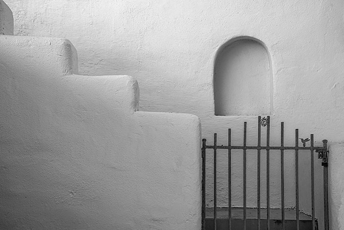 A minimalist architectural fine art print featuring a stepped wall, a small arched niche, and a simple iron gate, all rendered in shades of gray. The composition highlights the interplay between geometric shapes and textures.